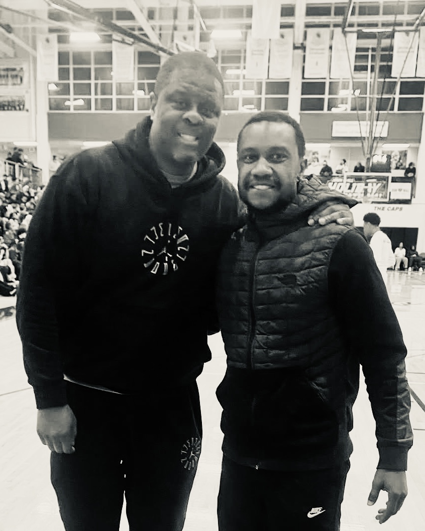 Coach Antonio Hicks with Coach Brian L. Clifton courtside in a packed gym, black and white.